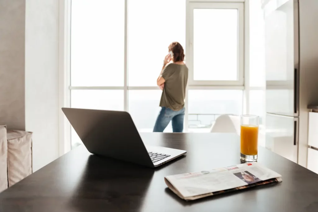 Eine telefonierende Frau im Homeoffice, auf dem Tisch ein Laptop, eine Zeitung und ein Glas. Hybride Arbeitsmodelle sind aktuelle Herausforderungen von Personalabteilungen.