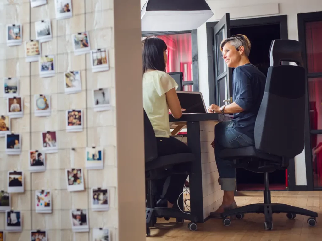 Zwei Frauen sitzen sich in einem modernen Büro an einem Schreibtisch gegenüber und führen ein Gespräch. Im Vordergrund ist eine Wand mit vielen Polaroid-Fotos zu sehen. Beide Frauen arbeiten im Jobsharing Modell an einem Laptop, das Büro wirkt hell und kreativ eingerichtet.
