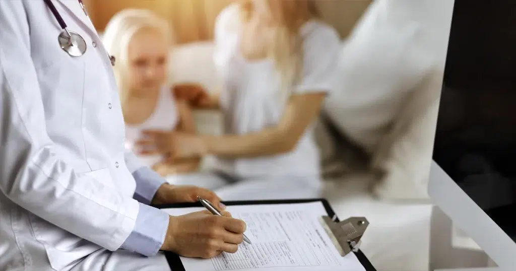 Krankenversicherung: Leistungen, Beiträge und Absicherung im Überblick Krankenversicherung: Ärztin dokumentiert Behandlung während Patientin mit Kind im Hintergrund