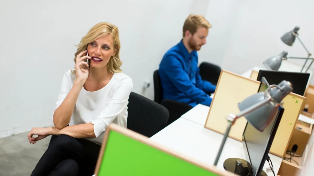 Eine Frau telefoniert an einem Desk Sharing Arbeitsplatz. Im Hintergrund arbeitet ein Mann konzentriert an seinem Computer.