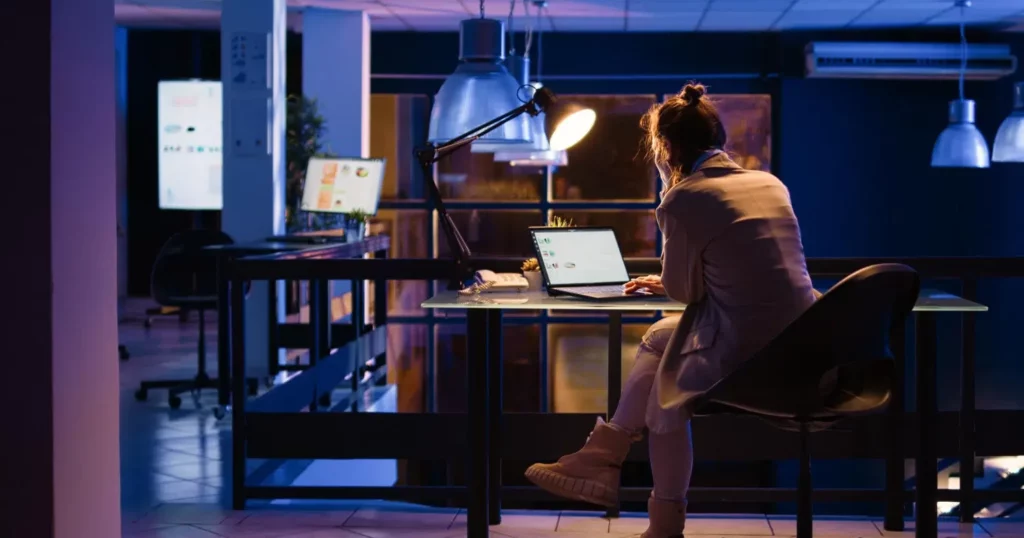 Eine Frau sitzt im Büro an einem Schreibtisch am Laptop. Nur eine Schreibtischlampe ist an, sonst ist alles dunkel. So kann eine Nachtschicht inkl. Nachtzuschlag aussehen.