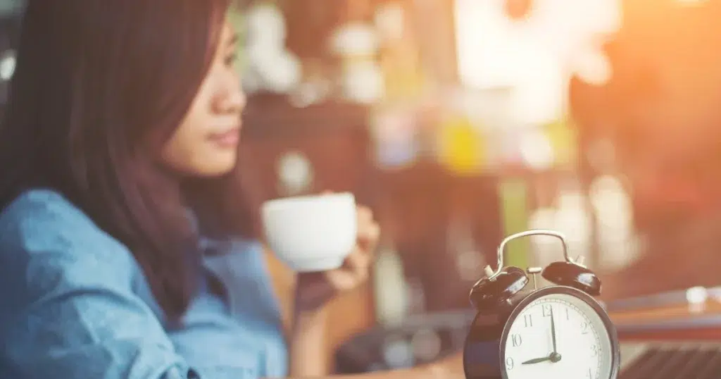 Frau macht Pause mit Kaffeetasse, Wecker auf dem Tisch – gesetzliche Pausenzeit im Arbeitsalltag