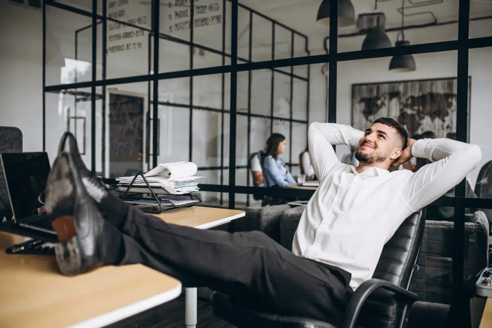 Formen von Arbeitszeitbetrug während der Arbeitszeit Mitarbeiter lehnt sich entspannt am Schreibtisch zurück während der Arbeitszeit und macht eine ausgiebige Pause